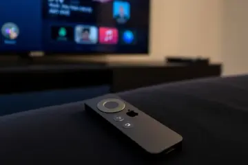 alt_text: A sleek Apple TV remote on dark fabric, with a modern IPTV app's interface reflected on the black TV screen behind it.